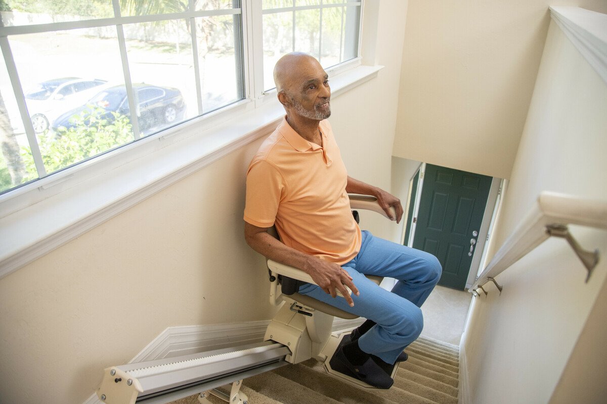 Senior man rides a straight-track stairlift down carpeted stairs in a bright home entryway, smiling.
