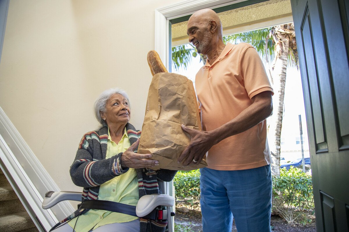 Stairlift with straight track at entryway; an elderly woman seated in the chair receives a grocery bag from a man.