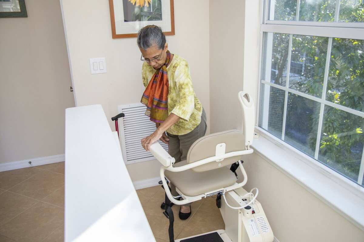 Elderly woman with cane riding a straight-track stairlift in a bright home hallway.