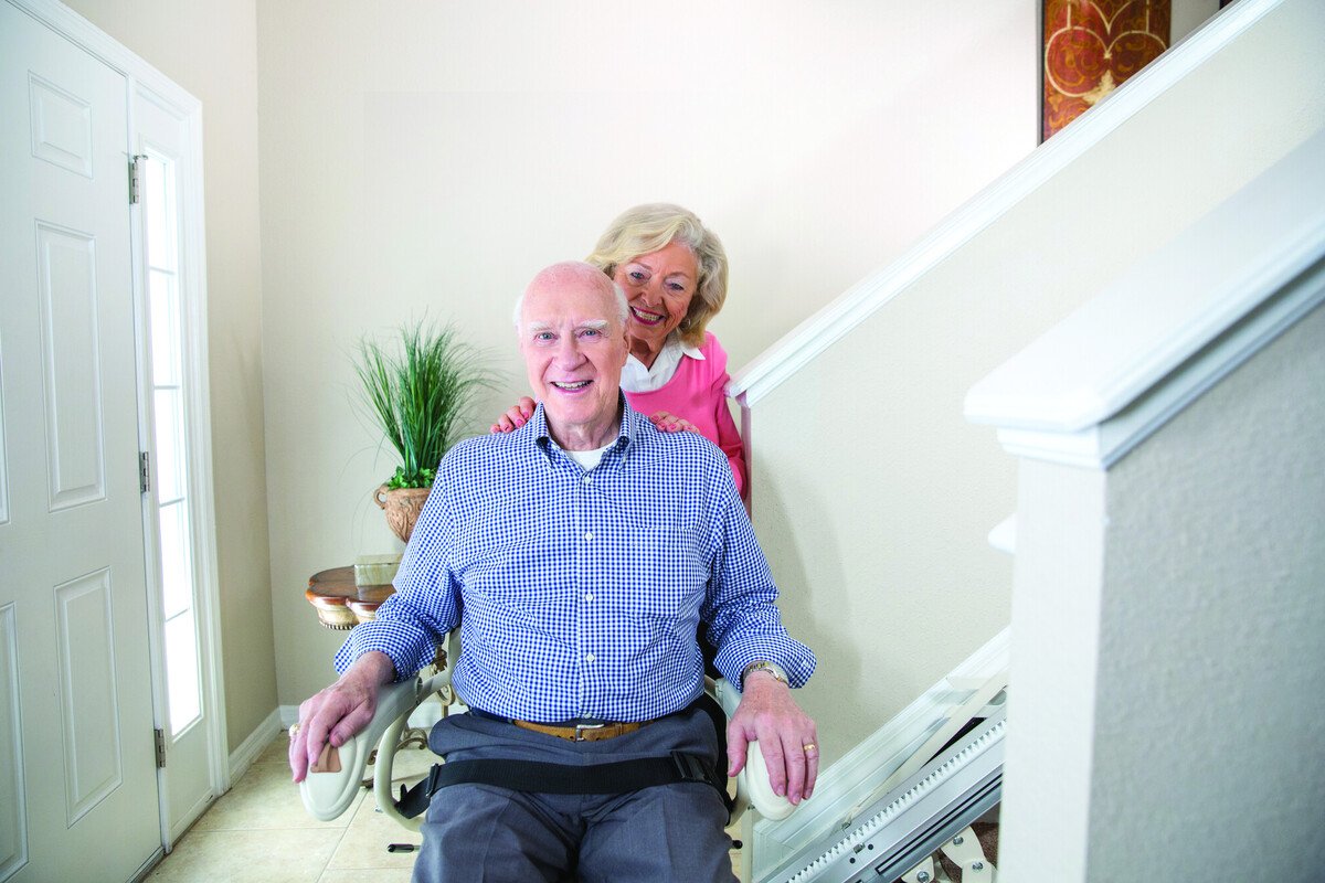 Elderly couple smiling at the bottom of a staircase on a stairlift, with the woman standing behind the seated man.